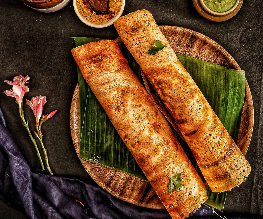 Two crisp golden dosai on a banana leaf on a wooden tray, with bowls of coconut chutney, green chutney and podi beside them.