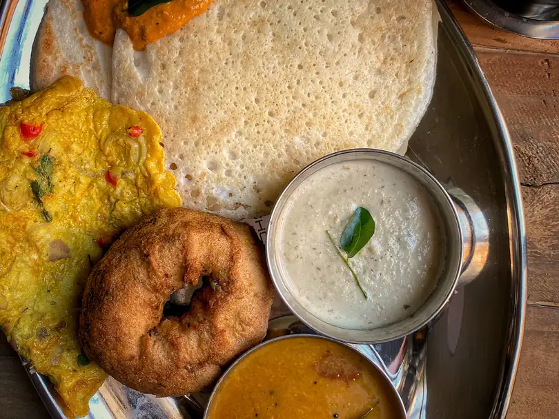 A steel plate with a soft set dosai topped with tomato chutney, rava kichadi, a medu vadai, coconut chutney and sambar, with filter coffee beside it.